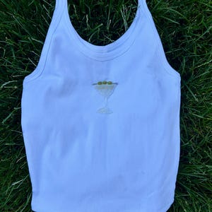 May include: A light blue ribbed tank top with spaghetti straps. The tank top features an embroidered martini glass design with green olives. The garment is made of a soft, lightweight material.
