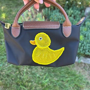 May include: Black handbag with a yellow beaded duck design. The bag has a brown leather handle and flap with white stitching. The zipper is gold. The bag is held against a background of green grass and foliage.