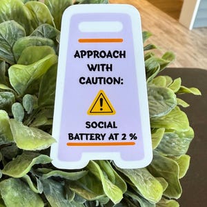 May include: A white and purple sign with the text "APPROACH WITH CAUTION: SOCIAL BATTERY AT 2 %" and a yellow warning symbol. The sign is placed in front of green foliage.