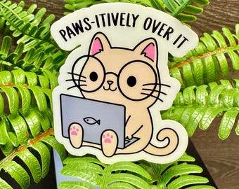 Könnte beinhalten: Ein Aufkleber mit einer Cartoon-Katze mit Brille, die an einem Laptop arbeitet. Die Katze ist hellbraun mit rosa Ohren und Pfoten. Der Laptop zeigt ein Fischsymbol. Der Text "PAWS-ITIVELY OVER IT" ist über der Katze.