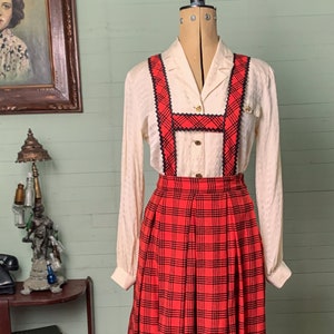 Pode incluir: Conjunto vintage de saia xadrez vermelha com detalhes em preto e suspensórios, combinado com uma blusa creme. A saia apresenta um design plissado e acabamento preto. O conjunto é exibido em um manequim, estilo retrô.