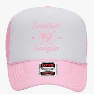 May include: A pink and white trucker hat with the words "Sensitive Gangsta" in pink script. The hat features a pink brim and mesh back, with a heart and arrow graphic. The hat has a snap closure.