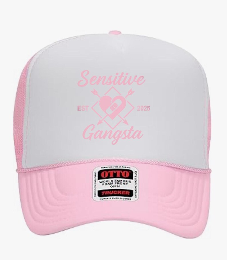 May include: A pink and white trucker hat with the words "Sensitive Gangsta" in pink script. The hat features a pink brim and mesh back, with a heart and arrow graphic. The hat has a snap closure.