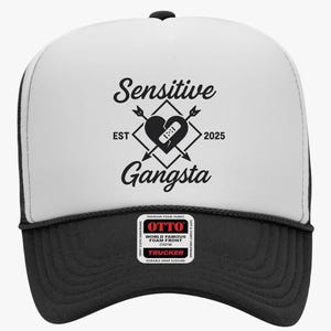 May include: A black and white trucker hat with the words "Sensitive Gangsta" and a heart graphic. The hat has a black brim and a white front panel with the text and graphic. The hat also has a black mesh back.