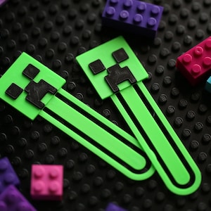 May include: Two green and black Minecraft Creeper-themed bookmarks are displayed with colourful building blocks. The bookmarks have a rectangular head and a long, thin body. The blocks are in shades of purple, pink, and blue.