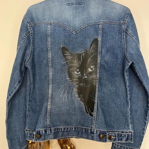 May include: A blue denim jacket featuring a detailed hand-painted illustration of a black cat on the back panel. The jacket has a classic collar, long sleeves, buttoned cuffs, and a button-down front closure with visible stitching and metal buttons.