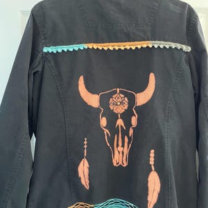May include: Black jacket with a collared neckline and button closure. The back features a peach-coloured bull skull design with feathers. The jacket is trimmed with colourful crochet lace in shades of turquoise, orange, and grey.