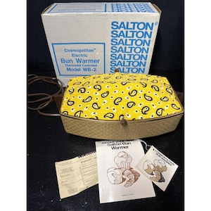 May include: A Salton Cosmopolitan electric bun warmer model WB-2 with a yellow and black paisley fabric lid and a woven-texture base. The set includes the original box, a warranty card, an instruction manual, and a product tag with illustrations.