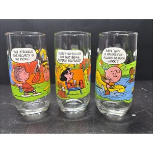 May include: Three clear glass tumblers featuring Peanuts comic strip illustrations. The first shows Linus with the text THE STRUGGLE FOR SECURITY IS NO PICNIC. The second shows Lucy with THERE'S NO EXCUSE FOR NOT BEING PROPERLY PREPARED. The third shows Charlie Brown with RATS! WHY IS HAVING FUN ALWAYS SO MUCH WORK?