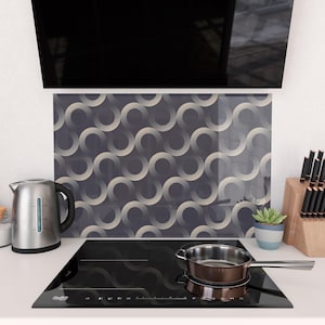 May include: A modern kitchen scene featuring a black stovetop with a stainless steel pan. A decorative backsplash with a repeating wave pattern in gray and beige tones is visible. A stainless steel kettle and a small potted plant are also present.