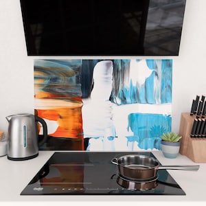 May include: A modern kitchen scene featuring a black stovetop with a stainless steel pan. A silver electric kettle and a colorful abstract art backsplash add to the contemporary design. A small succulent in a pot and a knife block are also visible.