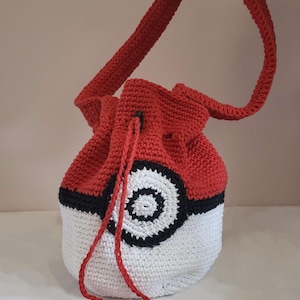 May include: A handmade crochet bag in the style of a Poké Ball. The bag is red and white with a black stripe and a circular design. It has a red drawstring closure and a red strap for carrying.
