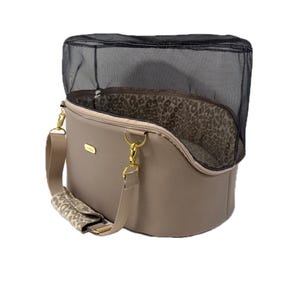 May include: A taupe pet carrier with a black mesh top and a leopard print interior lining. The carrier features gold-toned hardware, a matching taupe shoulder strap with a leopard print shoulder pad, and a small gold rectangular logo plate on the front.