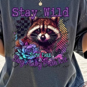 May include: Dark gray t-shirt featuring a raccoon graphic with the text "Stay Wild, Stay Free & Stay Away From Me." The design includes floral elements and a checkered pattern. The shirt has a relaxed fit.