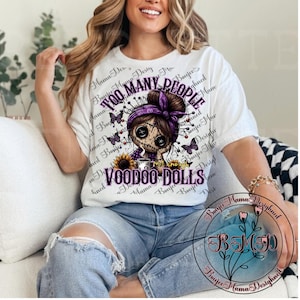 May include: White t-shirt featuring a voodoo doll design with the text "TOO MANY PEOPLE" and "VOODOO DOLLS". The graphic includes a doll with a purple headscarf, butterflies, and a sunflower. The shirt is worn by a person.
