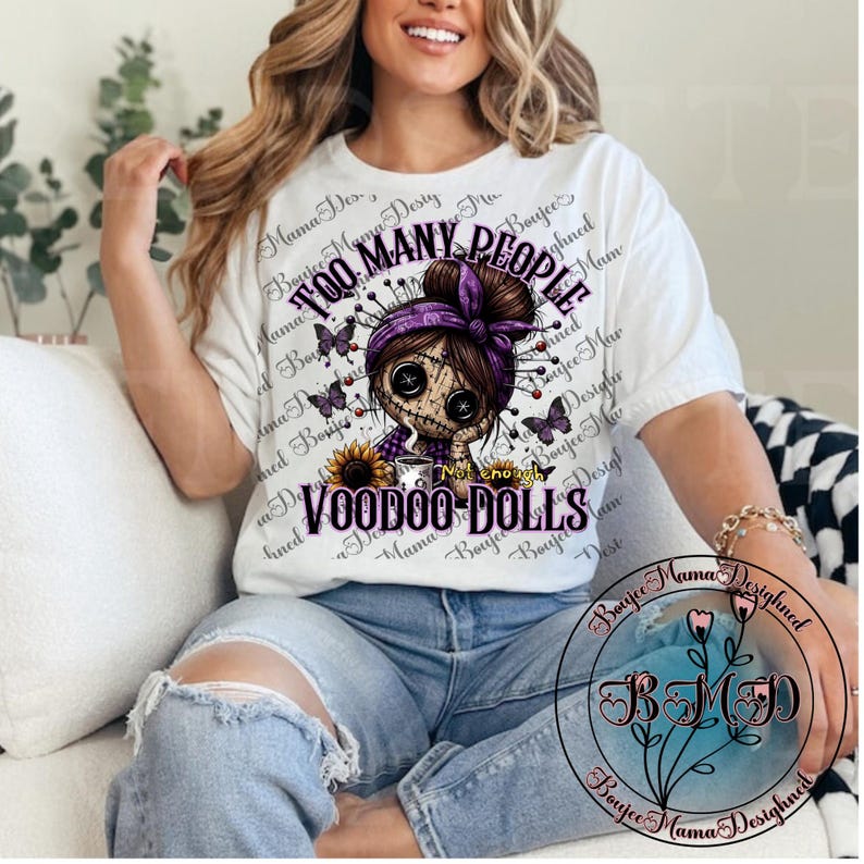 May include: White t-shirt featuring a voodoo doll design with the text "TOO MANY PEOPLE" and "VOODOO DOLLS". The graphic includes a doll with a purple headscarf, butterflies, and a sunflower. The shirt is worn by a person.