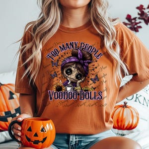May include: A burnt orange t-shirt with the text "TOO MANY PEOPLE" and "VOODOO DOLLS". The graphic features a voodoo doll with a purple headscarf, surrounded by butterflies and text. A pumpkin-shaped mug is held in the foreground.