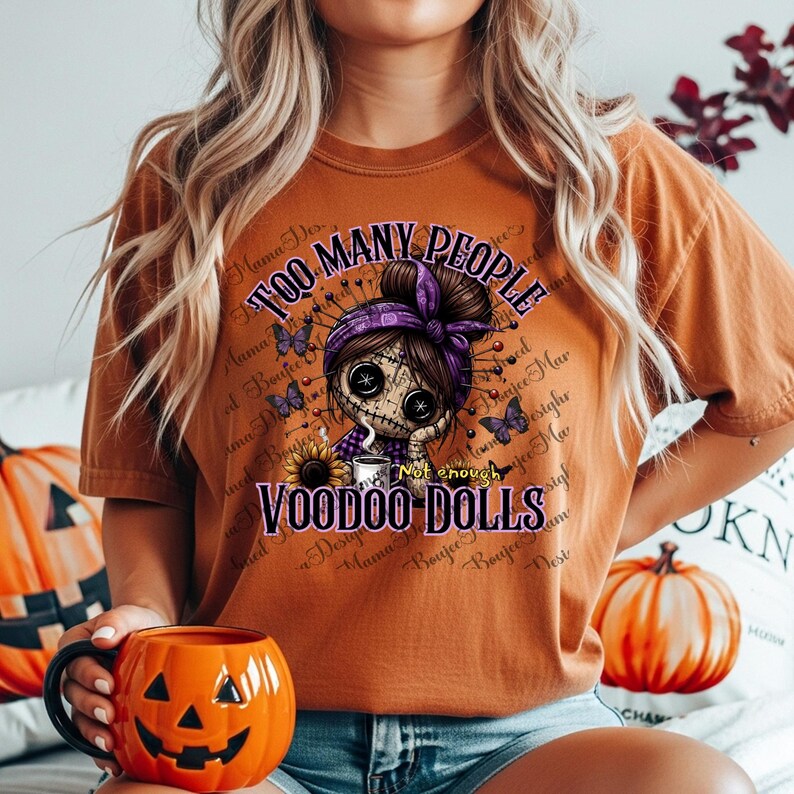 May include: A burnt orange t-shirt with the text "TOO MANY PEOPLE" and "VOODOO DOLLS". The graphic features a voodoo doll with a purple headscarf, surrounded by butterflies and text. A pumpkin-shaped mug is held in the foreground.