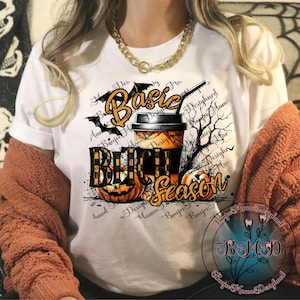 Könnte beinhalten: Weißes T-Shirt mit einem Halloween-Motiv. Das Design zeigt die Worte "Basic Bitch Season" in Orange und Schwarz, einen Kaffeebecher, Fledermäuse und Kürbisse. Das T-Shirt hat kurze Ärmel.