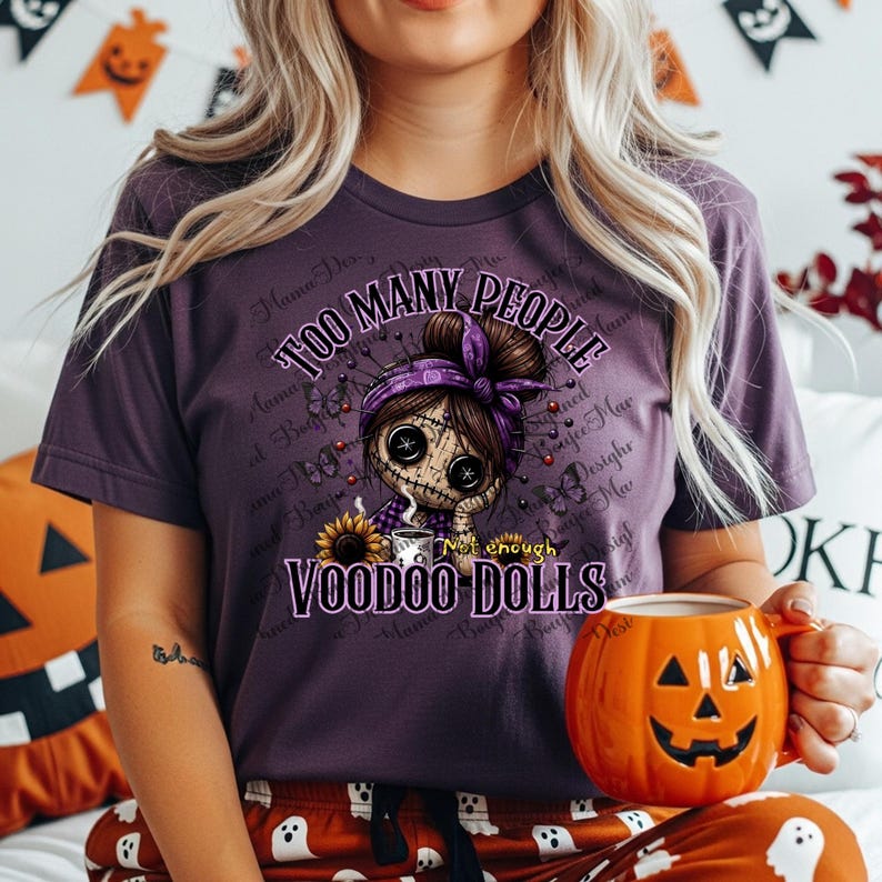 May include: Purple t-shirt with the text "TOO MANY PEOPLE, NOT ENOUGH VOODOO DOLLS" and a graphic of a voodoo doll. The doll has button eyes and is wearing a purple headband. The person is holding an orange pumpkin mug.