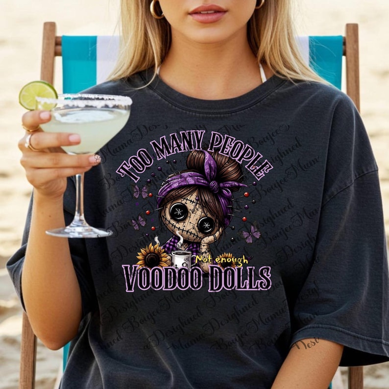 May include: Dark gray t-shirt with the text "TOO MANY PEOPLE, VOODOO DOLLS" and a graphic of a voodoo doll with a purple headband, holding a cup of coffee. The doll is surrounded by butterflies and a sunflower.