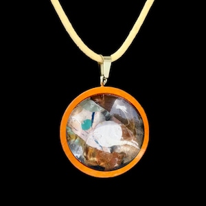 May include: A round pendant necklace with an orange frame and a clear center filled with colorful abstract art. The pendant hangs from a light yellow cord. The background is black.