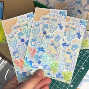 Peut inclure: Trois planches d'autocollants avec un motif de "récif corallien". Les autocollants présentent des illustrations de créatures marines, de coraux et de bulles en bleu, rose et vert sur un fond vert clair. Le texte "coral reef" est imprimé en haut.