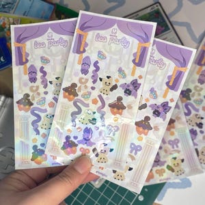 Peut inclure: Trois planches d'autocollants à finition holographique, sur le thème de la "Spooky Tea Party". Les autocollants comprennent des personnages de dessins animés, des cupcakes, des tasses à thé et des éléments décoratifs violets, marron et blancs.