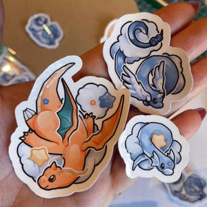 Peut inclure: Plusieurs autocollants colorés représentant des dragons de dessins animés. On y voit un dragon orange avec des ailes turquoise et deux dragons bleus, le tout entouré de nuages et d'étoiles. Les autocollants ont un bord blanc.