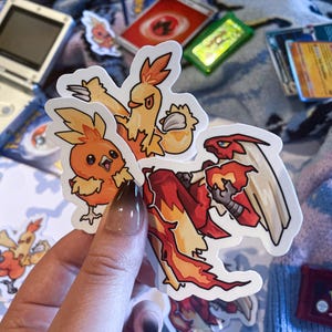 Puede incluir: Una colección de pegatinas de Pokémon coloridas con Torchic, Combusken y Blaziken. Las pegatinas se sostienen frente a un fondo con una Game Boy Advance SP, cartas de Pokémon y un cartucho de Pokémon Rubí.
