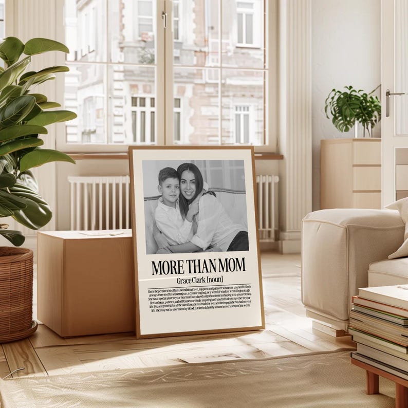 May include: A framed black and white print with the text "MORE THAN MOM" and a photo of a woman and child. The print is in a light brown frame and is displayed in a well-lit room with a large window and a plant.