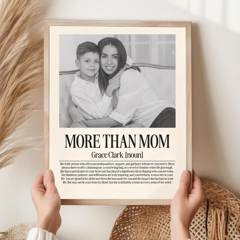 May include: A framed print with a black and white photo of a woman and child. The text "MORE THAN MOM" is displayed below the photo, with the name "Grace Clark" and a definition. The print is held by hands.