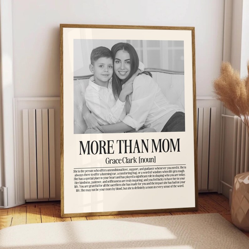 May include: A framed black and white print featuring a mother and child. The text "MORE THAN MOM" is displayed below the image, with additional text describing a mother's role. The frame is a warm, natural wood tone.