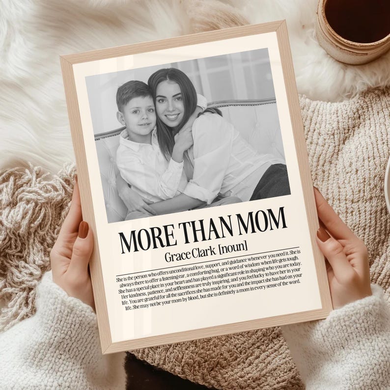 May include: A framed print with the text "MORE THAN MOM" and a black and white photo of a mother and child. The print has a light wood frame and is held by a person wearing a cream-colored sweater.