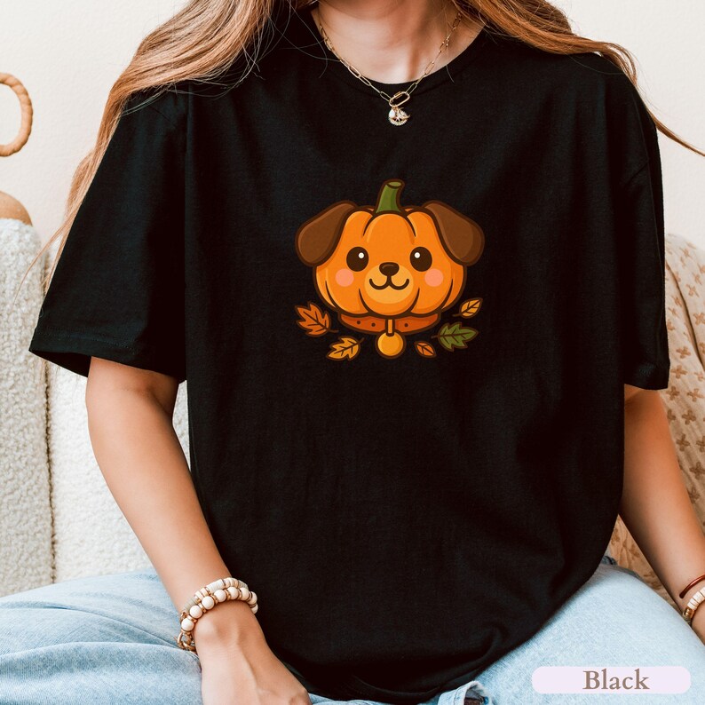May include: Black t-shirt with a cartoon dog-pumpkin design. The dog has brown ears and a pumpkin-shaped face with a smiling expression. The design includes a brown collar and autumn leaves. The word "Black" is at the bottom.