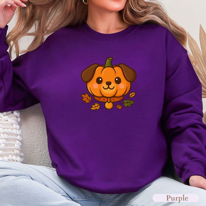 May include: A purple sweatshirt with a cartoon dog-pumpkin design. The dog has brown ears and a collar with a gold tag. Autumn leaves surround the dog. The word "Purple" is at the bottom of the image.