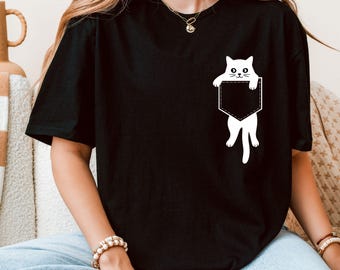 May include: Black t-shirt featuring a white cat graphic peeking out of a pocket design. The cat has a simple, cartoon-like style. The word "Black" is printed at the bottom of the image. The shirt is a casual style.