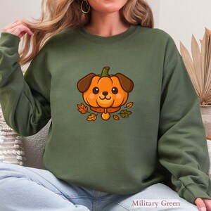 May include: Military green sweatshirt with a cartoon pumpkin-dog design. The orange pumpkin-dog has brown ears and a collar, surrounded by autumn leaves. The text "Military Green" is at the bottom right.