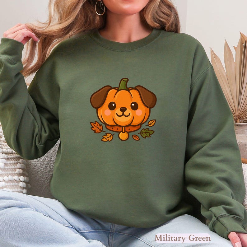 May include: Military green sweatshirt with a cartoon pumpkin-dog design. The orange pumpkin-dog has brown ears and a collar, surrounded by autumn leaves. The text "Military Green" is at the bottom right.