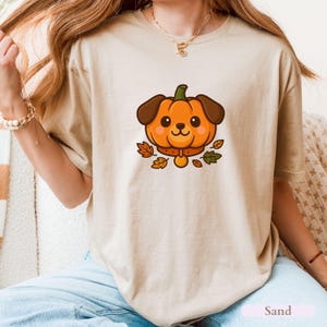 May include: Sand-coloured t-shirt featuring a cartoon dog with a pumpkin head. The dog has brown ears and a collar, surrounded by autumn leaves. The shirt is a neutral colour, perfect for autumn.