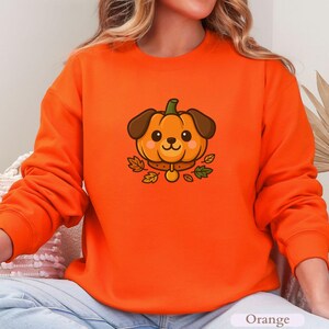 May include: Bright orange sweatshirt with a cartoon dog design. The dog has a pumpkin-shaped head, brown ears, and a gold collar. Green and brown leaves surround the dog. The word "Orange" is printed at the bottom.
