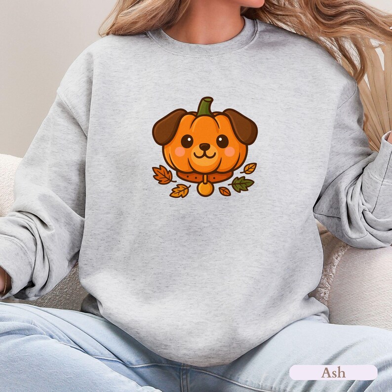 May include: A light grey sweatshirt with a cartoon dog designed as a pumpkin. The dog has brown ears and a collar, surrounded by autumn leaves. The word "Ash" is printed at the bottom.