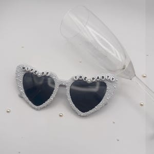 K&ouml;nnte beinhalten: Herzf&ouml;rmige Sonnenbrille mit schwarzen Gl&auml;sern und wei&szlig;en Perlen. Auf der Brille steht in schwarzen Buchstaben "WIFE OF THE PARTY". Ein Sektglas liegt hinter der Brille auf wei&szlig;em Hintergrund.