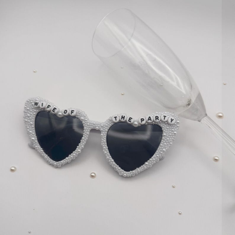 K&ouml;nnte beinhalten: Herzf&ouml;rmige Sonnenbrille mit schwarzen Gl&auml;sern und wei&szlig;en Perlen. Auf der Brille steht in schwarzen Buchstaben "WIFE OF THE PARTY". Ein Sektglas liegt hinter der Brille auf wei&szlig;em Hintergrund.