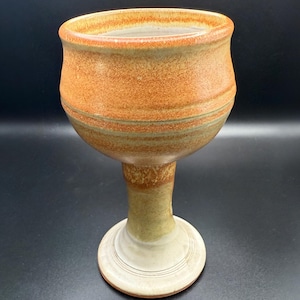 May include: A handmade ceramic goblet with a wide, textured orange bowl and a slender stem. The goblet has a round, off-white base with concentric rings. The stem is light green and brown, and the bowl has green and brown bands.