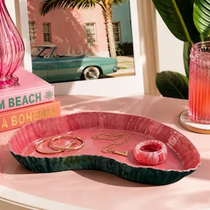 May include: An irregular, kidney-shaped ceramic tray with a glossy pink interior and a dark green, textured exterior. The tray holds two gold-toned metal bangles, a delicate gold chain necklace with a letter pendant, and a pink and white marbled ring.