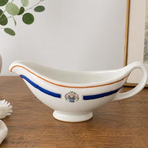 May include: A white ceramic gravy boat featuring a curved handle, a pedestal base, and a painted design consisting of a thin orange rim, a bold blue stripe, and a small central floral motif depicting a basket of pink and green flowers.