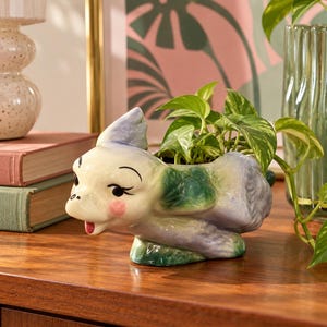May include: A ceramic planter shaped like a stylized animal with a glossy finish in shades of blue, green, and white. The animal figure features painted black eyes, pink cheeks, and a small red mouth, with a green plant growing from its back.