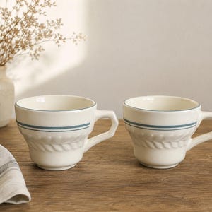 May include: Two cream-colored ceramic mugs featuring a distinct geometric handle and a horizontal blue stripe accent. The lower portion of each mug displays a raised, embossed rope-like texture encircling the base, providing a tactile detail.