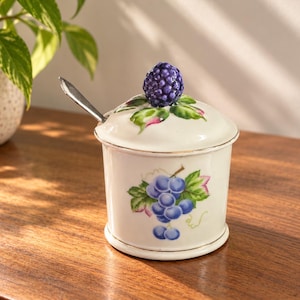 May include: A white ceramic jar with a lid featuring a sculpted purple blackberry handle. The jar is decorated with a painted cluster of blue grapes and green leaves. A silver-toned metal spoon is inserted into the side of the lid.